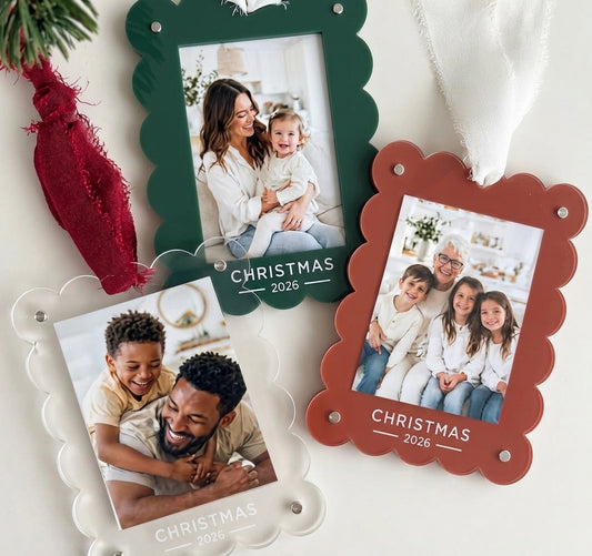 Three scalloped-edge acrylic Christmas photo ornaments in dark green, terracotta brown, and clear, each holding a family photo and the text 'CHRISTMAS 2026'. The ornaments are arranged on a white surface with holiday styling, including a red and white chiffon ribbon.