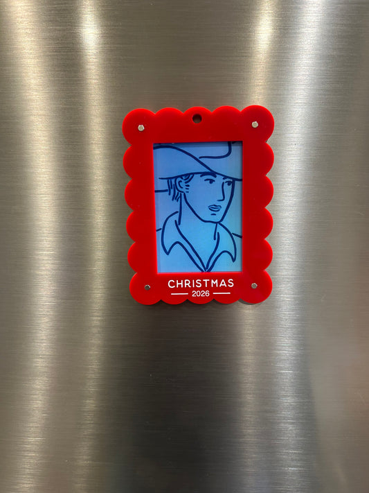 A red scalloped-edged Christmas 2026 magnetic photo holder with a blue-toned illustration of a cowboy in a hat, attached to a brushed stainless steel surface.